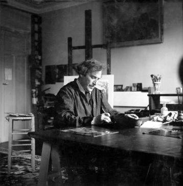 Marc Chagall en costume, écrit sur la table de l'atelier, devant l'œuvre Le Cimetière accrochée au mur.