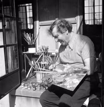 Marc Chagall smiling while using his color palette, with brushes and small containers behind him.