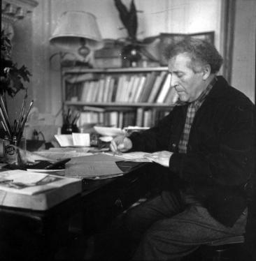 Marc Chagall écrit à son bureau, une bibliothèque et un pot de pinceaux en arrière-plan.