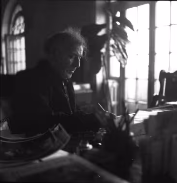 Marc Chagall writing at his desk, with a plant and two large windows in the background.