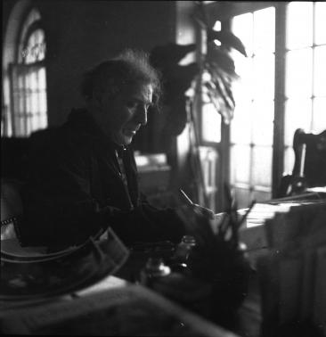 Marc Chagall écrit à son bureau, une plante et deux grandes fenêtres en arrière-plan.