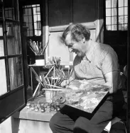 Marc Chagall smiling while using his color palette, with brushes and small containers behind him.