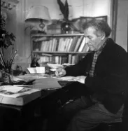 Marc Chagall writing at his desk, with a bookshelf and jar of brushes in the background.