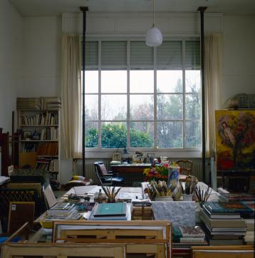 Color photograph of a tidy studio, with a window overlooking a garden and the painting The Branch in the background.