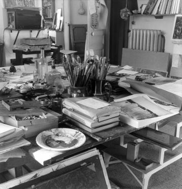 Large work surface covered with brushes, piles of books, printed images, and color palettes.