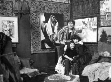 Marc and Bella Chagall, next to their daughter Ida, posing on a divan in their living room, with five paintings hanging behind them.