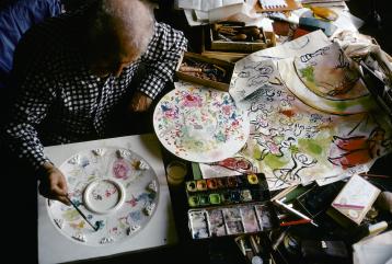 Chagall, cheveux blancs, travaillant sur une maquette pour l'Opéra de Paris à côté de palettes de couleurs et d'autres maquettes.