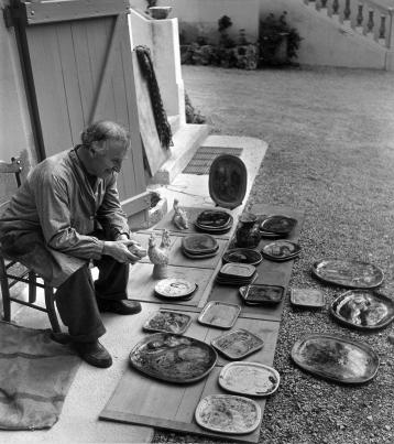 Marc Chagall, assis dans une cour intérieure, regarde une vingtaine de céramiques posées devant lui.