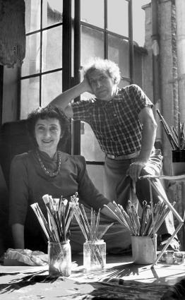 Marc Chagall standing and Bella Chagall sitting, smiling behind several jars filled with brushes.