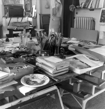 Vaste plan de travail recouvert de pinceaux, piles de livres, images imprimées et palettes de couleur.