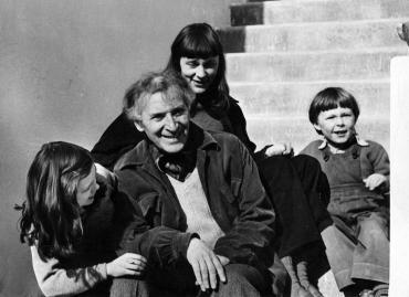 An elderly Marc Chagall smiling between two children (one of which is his son), sitting on a staircase. His partner is behind them.