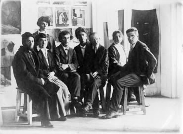 Photograph of a group of professors (including several women) sitting on wooden stools, in front of works on paper.
