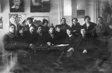 Photographie de groupe de professeurs chaudement habillés, Chagall au centre, dans une salle aux murs recouverts de tableaux.