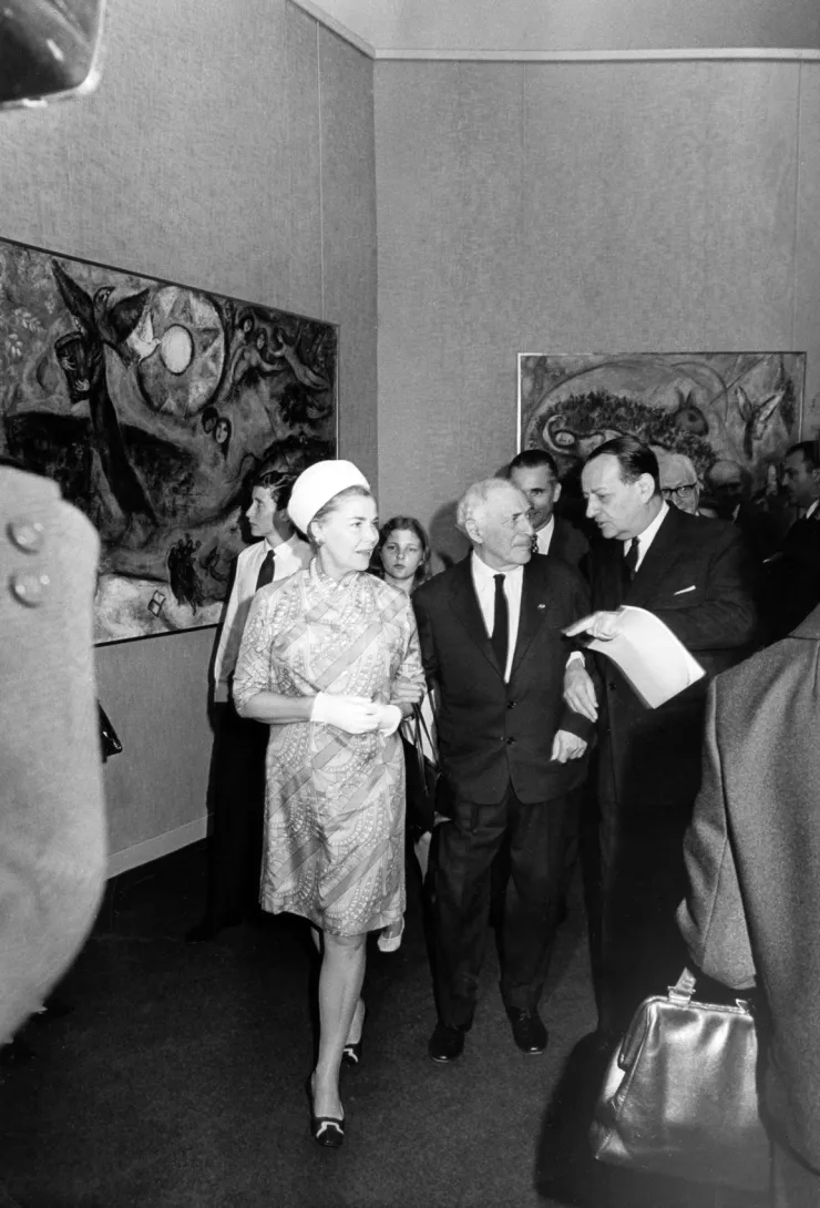 Marc Chagall et Madame de Vilmorin écoutent André Malraux dans une salle d'exposition, 2 tableaux de Chagall en arrière-plan.