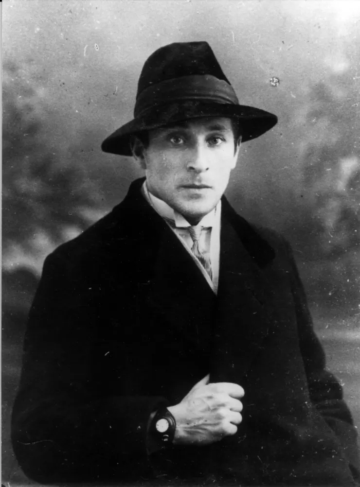 Chagall, jeune homme, porte un chapeau, un pardessus et une montre de couleur sombre.